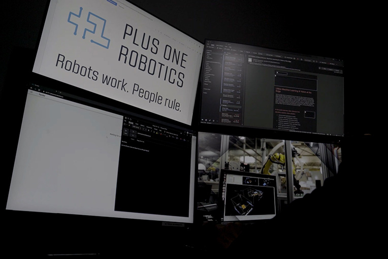 A man overseeing the work of robots in an office at a 4 monitor workstation